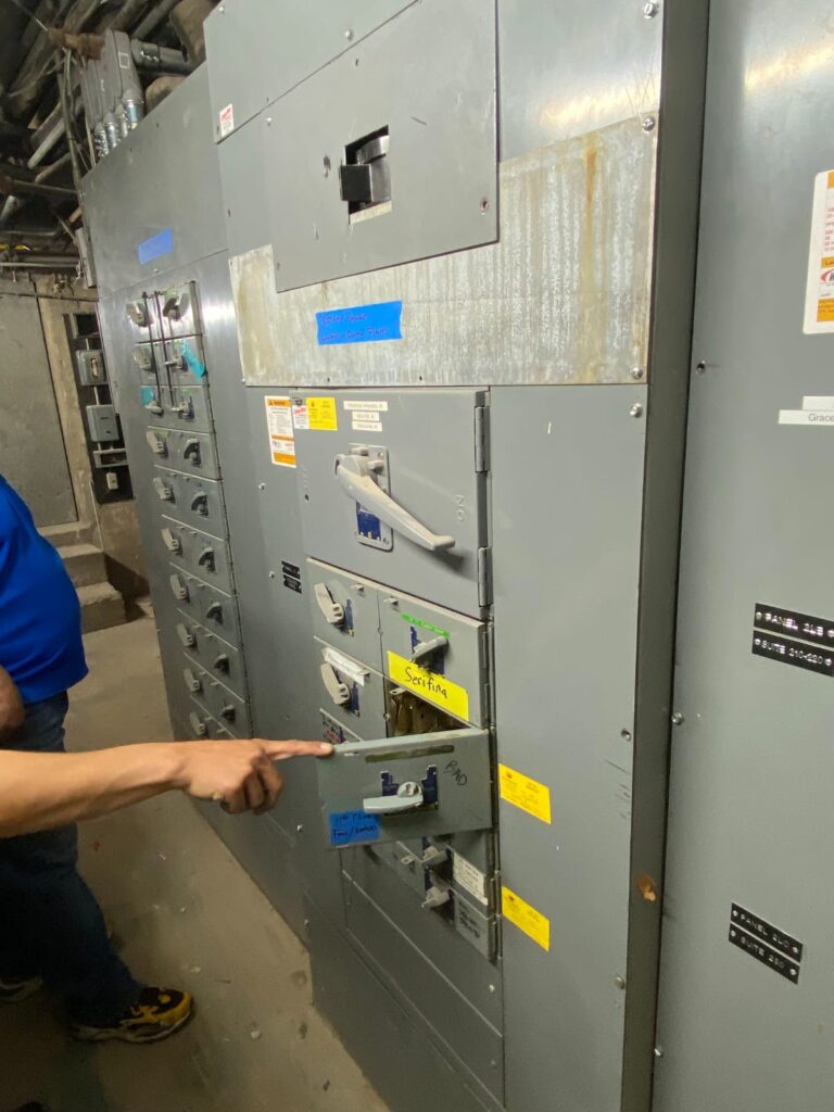 Electrician pointing to a labeled breaker on a commercial electrical switchboard